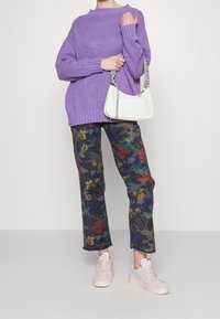 Personne portant un pull en tricot violet, un pantalon camouflage à motifs floraux, des baskets rose clair et tenant un sac à main blanc avec une bandoulière tressée.