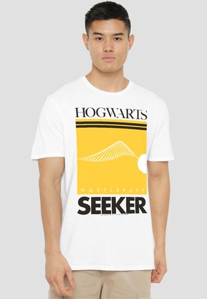 Young man wearing a white t-shirt with yellow and black Hogwarts Hufflepuff Seeker graphic, standing against a plain light gray background.