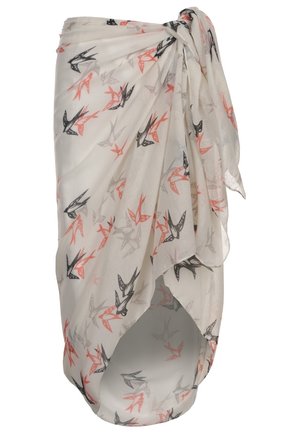 Femme portant un haut de bikini rouge et blanc à pois et un paréo blanc à imprimé d’oiseaux noué à la taille, debout pieds nus sur un fond uni.
