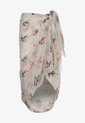 Femme portant un haut de bikini rouge et blanc à pois et un paréo blanc à imprimé d’oiseaux noué à la taille, debout pieds nus sur un fond uni.