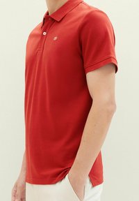 Rød polo-shirt med klassisk krave og korte ærmer. Den har tre knapper, sideskår og et diskret logo på venstre bryst samt tekstureret stof.