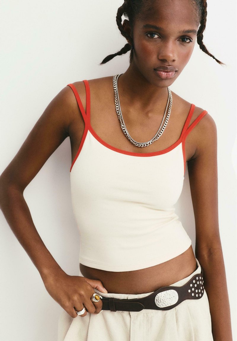 Jonge vrouw met gevlochten haar, gekleed in een witte crop top met rode schouderbanden, zilveren kettingen, ringen en beige broek met een brede bruine riem.