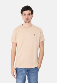 T-shirt beige in cotone con maniche corte, scollo rotondo e piccolo logo rosso sul petto. Indossato con jeans blu. Texture liscia.