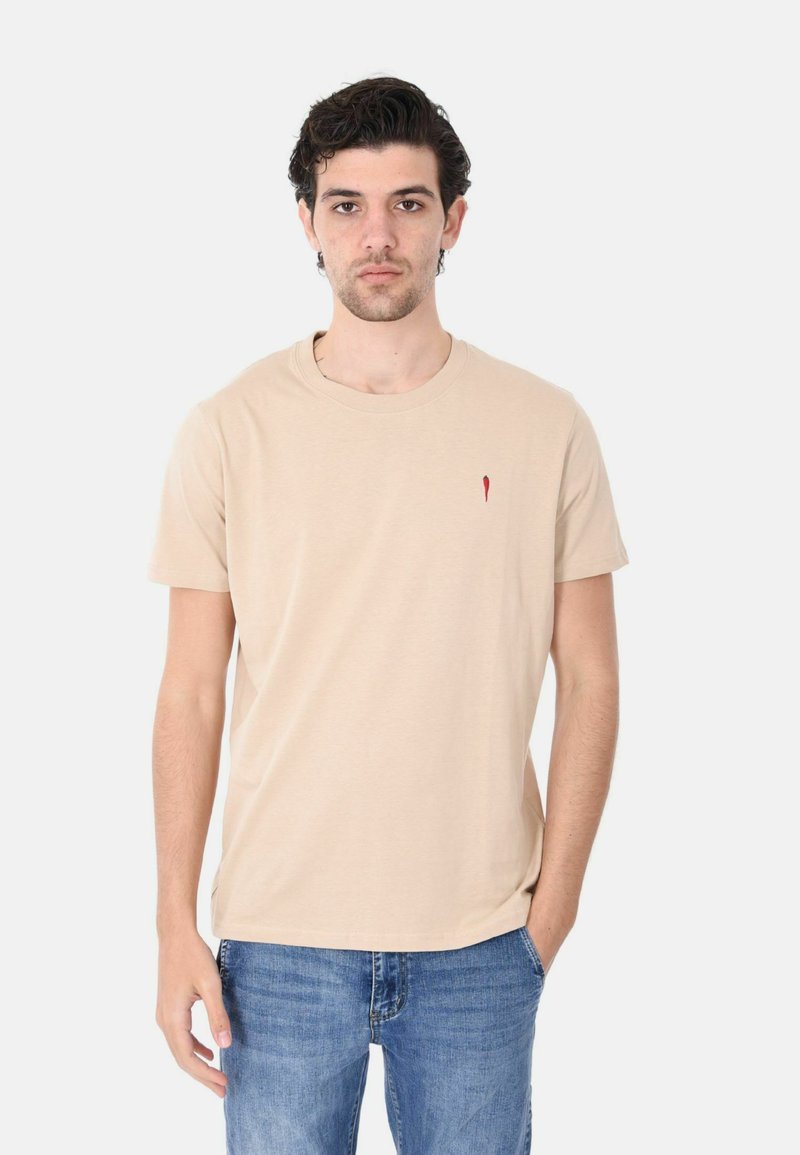 T-shirt beige in cotone con maniche corte, scollo rotondo e piccolo logo rosso sul petto. Indossato con jeans blu. Texture liscia.