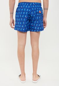 Pantaloncini da bagno blu con un motivo ripetuto della parola "SUNDEK" in lettere grasse. Vita elastica, tasca sul retro con chiusura a bottone.