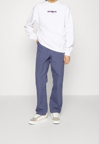 Sudadera blanca con texto morado, combinada con pantalones azules holgados y zapatillas beige con acentos blancos. Diseño simple y casual.