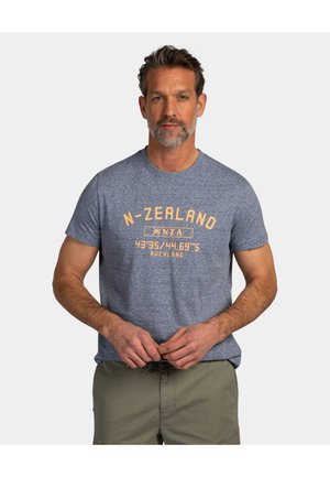 Man met kort haar en baard, die een blauw T-shirt met een Nieuw-Zeelandse afbeelding draagt en olijfgroene broek, staand tegen een effen achtergrond.