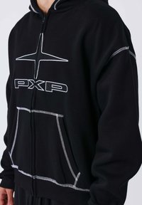 Sudadera con capucha negra y costuras blancas. Cuenta con una cremallera frontal, grandes bolsillos frontales y un logotipo estilizado en el pecho.