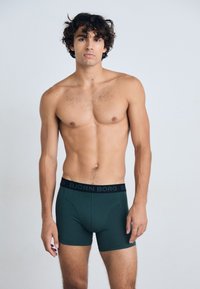Boxer scuri verdi con elastico nero in vita che presenta il logo "BJÖRN BORG", texture liscia e design aderente, indossati da un modello a petto nudo.