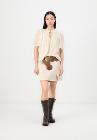 Krembluse med puffermer og knytedetalj, parret med en beige skjørt med en slits og brunt lærbelte med sirkulære detaljer. Brune knelang støvler.