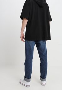 Person klädd i en svart överdimensionerad huva, uppkavlade blå jeans och vita högtopps Converse-sneakers, står mot en plain vit bakgrund.