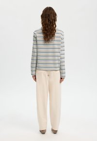 Μπλούζα με μακριά μανίκια και ριγέ σχέδιο σε κρεμ, μπλε και πράσινο. Συνδυάζεται με μπεζ παντελόνι φαρδιάς γραμμής. Το μοντέλο κοιτάζει προς τα πίσω, δείχνοντας το σχέδιο στο πίσω μέρος του ρούχου.