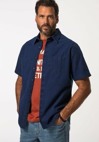 Camicetta a maniche corte di colore blu navy con chiusura a bottoni e due tasche sul petto, indossata sopra una maglietta grafica rossa, abbinata a jeans blu chiaro.