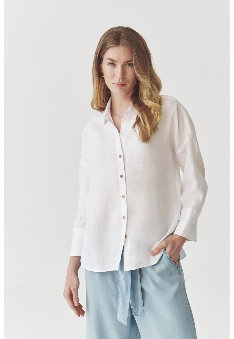 White linen button-up shirt with long sleeves, slightly oversized fit, featuring light brown buttons and a pocket on the left side.