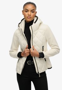 Biała pikowana kurtka z kapturem, czarnymi akcentami i zamkami. Materiał wydaje się miękki, ma dopasowany krój i teksturowany wzór.