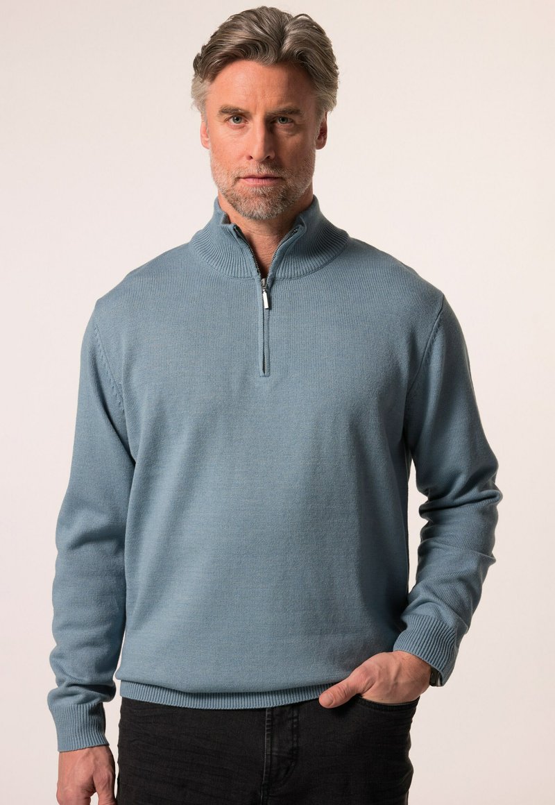 Světlomodrý pletený svetr s vysokým límcem, s polovinou zipu vpředu, žebrovanými manžetami a hladkou texturou. Nosený přes černé kalhoty.