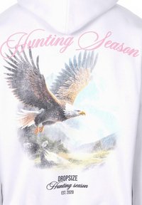 Hoodie blanc présentant un graphique détaillé d'un aigle en plein vol, avec le texte "Saison de chasse" en rose et "Dropsize EST. 2020" en dessous.