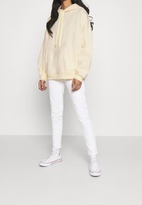 Felpa gialla chiara con cappuccio regolabile, grande tasca frontale e polsini a costine, abbinata a jeans bianchi slim-fit e sneaker bianche.