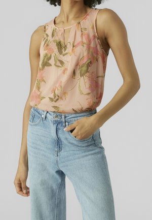 Ærmeløs ferskenfarvet bluse med blomsterprint, stoppet ned i lyseblå, højtaljede jeans, med den ene hånd i lommen og neutral baggrund.