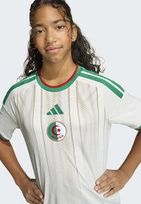 Maillot de football blanc avec des accents verts et rouges, présentant des rayures verticales et un écusson du drapeau algérien. Logo Adidas sur l'épaule.