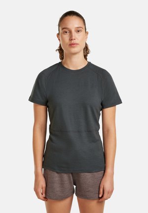 Kvinde med brunt hår sat op, iført en mørkegrå T-shirt med korte ærmer og brune shorts, stående foran en helt hvid baggrund.