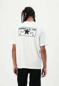 Baltas kokvilnas T-krekls ar lielu melnu grafiku uz muguras, kurā redzams teksts "CONVERSE ALL STAR" un centrālais zvaigznes dizains. Īsas piedurknes.