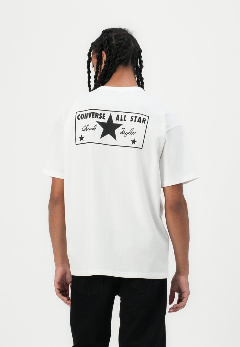 Baltas kokvilnas T-krekls ar lielu melnu grafiku uz muguras, kurā redzams teksts "CONVERSE ALL STAR" un centrālais zvaigznes dizains. Īsas piedurknes.