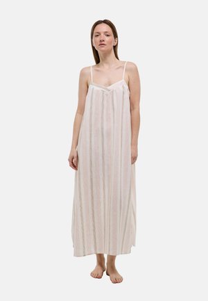 Femme debout pieds nus, portant une longue robe rose clair sans manches avec des rayures verticales beiges et blanches sur un fond uni.