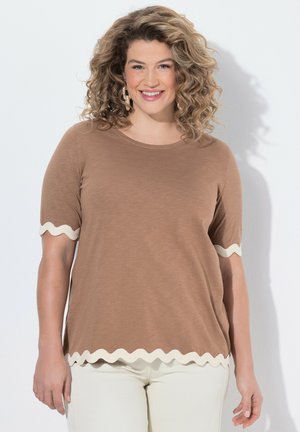 Lächelnde Frau mit lockigem Haar, die ein braunes kurzärmeliges Oberteil mit weißem wellenförmigem Saum und weiße Hose trägt, vor einem hellen Hintergrund stehend.