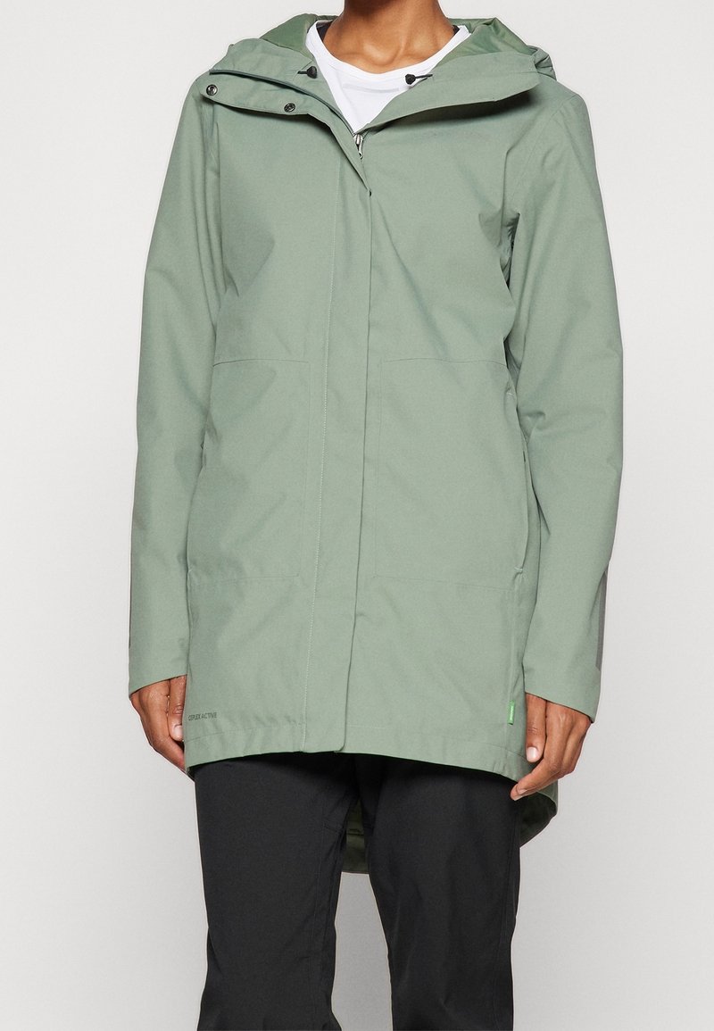 Personne portant un imperméable à capuche vert clair avec des poches avant par-dessus une chemise blanche et un pantalon noir, debout devant un fond uni.