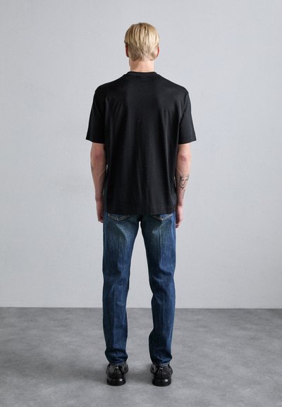 Juodas trumpas rankovėmis marškinėliai su laisvu siluetu, derinami su mėlynomis džinsomis, matomi iš nugaros. Marškiniai turi subtilų tekstūros raštą.