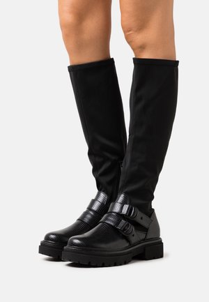 Botas negras hasta el tobillo con material texturizado, acentos de doble hebilla y suelas gruesas. Combinadas con cañones de tela negra que llegan hasta las pantorrillas.