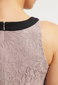 Koronkowa sukienka ma jasnoróżowy wzór w kwiaty z czarnym dekoltem i detalem w postaci zamka z tyłu. Tekstura jest miękka i skomplikowana.