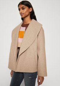Μπεζ παλτό από μείγμα μαλλιού με φαρδιές πέτες, ίσια γραμμή και μπροστινές τσέπες, σε συνδυασμό με πλεκτό πουλόβερ σε πολύχρωμες ρίγες και σκούρα τζιν.