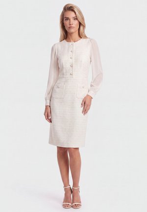 Blonde woman stands wearing a light pink knee-length dress with long sheer sleeves, front buttons, two waist pockets, and matching high-heeled sandals.