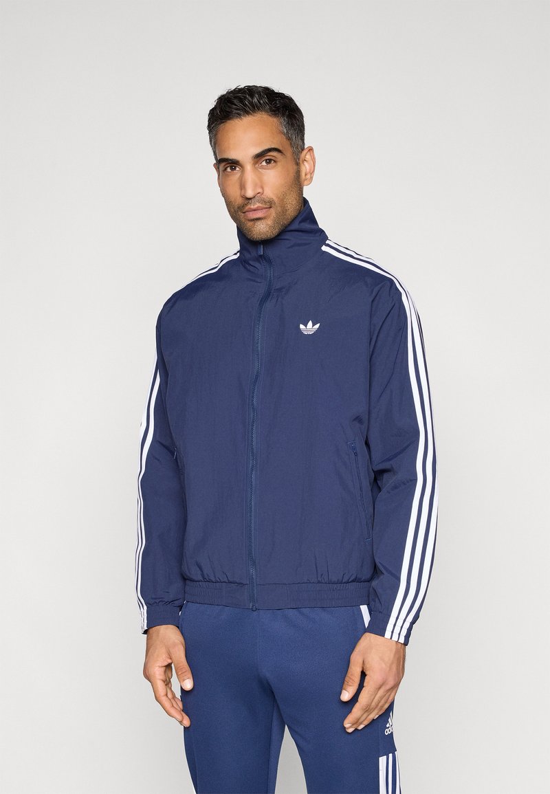 Vetrelecová vetrovka tmavomodrej farby s celým zipsom, vysokým golierom a bielymi pruhmi pozdĺž rukávov; na hrudi je logo Adidas.