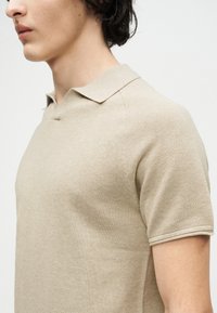 Πλάγια όψη προσώπου που φοράει μπεζ πλεκτό κοντομάνικο πόλο πουκάμισο με υφή υφάσματος σε απλό ανοιχτό φόντο.