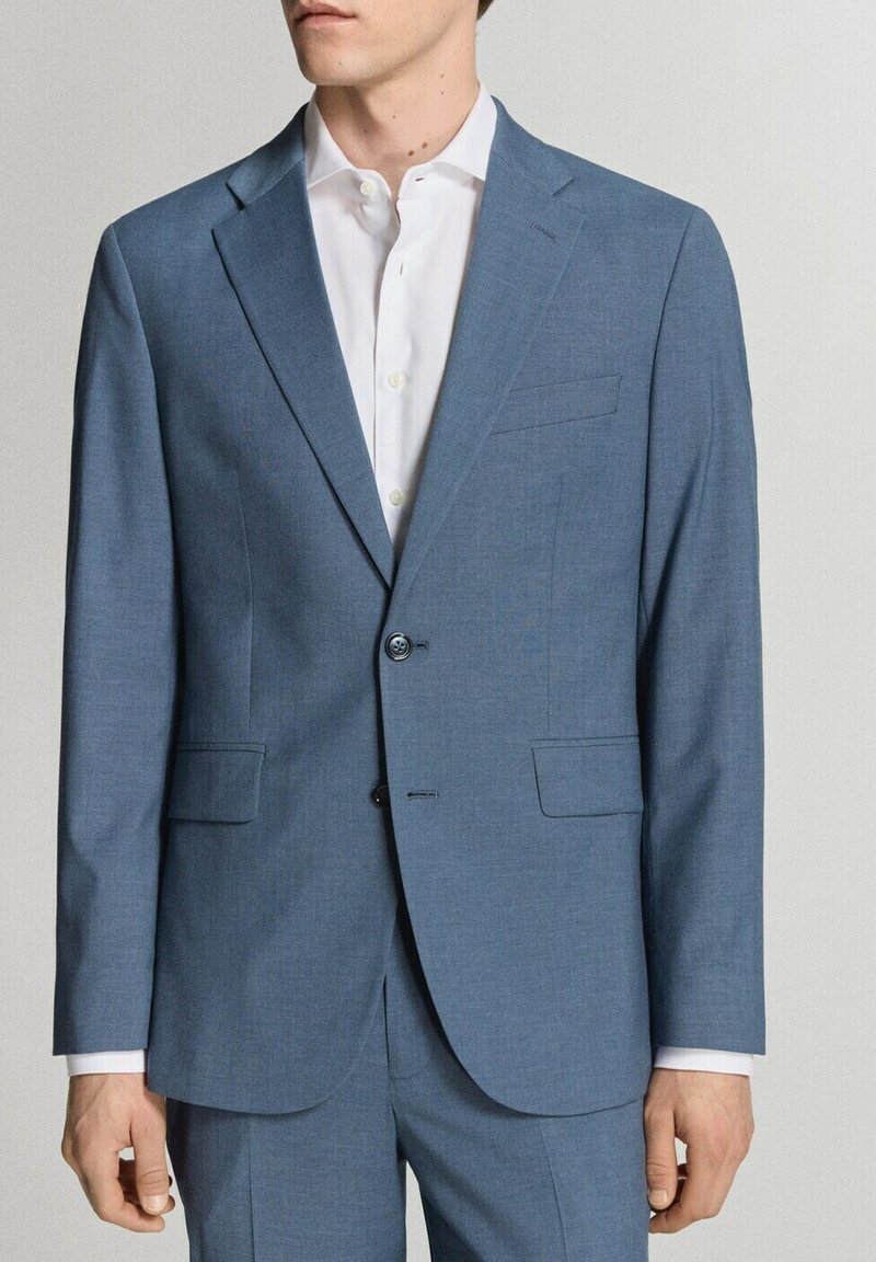 Hombre con chaqueta de traje azul claro de dos botones y pantalones a juego sobre una camisa blanca de vestir, de pie contra una pared lisa.