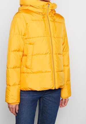 Kvinde iført en knaldgul quiltet dunjakke med lynlås foran, hætte med snore og blå jeans, stående mod en hvid baggrund.