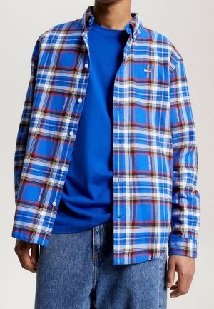 Man die een blauw geruit overhemd met knopen draagt over een effen blauw T-shirt met blauwe spijkerbroek, staand tegen een effen achtergrond.