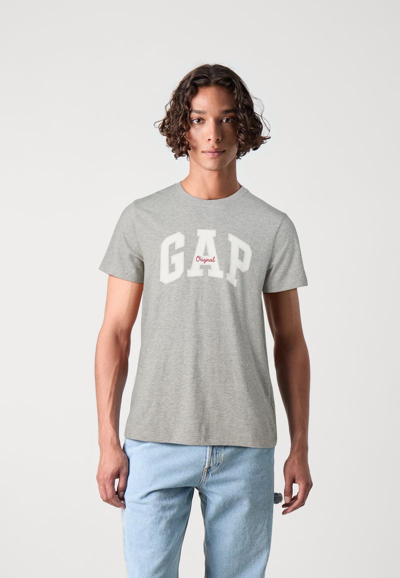 Tricou gri din bumbac cu litere albe îndrăznețe "GAP" și detaliu roșu "Original". Mâneci scurte, guler rotund, croială lejeră.