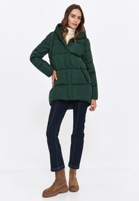 Ciemnozielona kurtka puchowa z kapturem, o pikowanej fakturze, z bocznymi kieszeniami i lekko zaokrąglonym dołem, w połączeniu z ciemnoniebieskimi dżinsami i brązowymi butami.