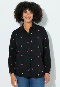 Vrouw met krullend haar die lacht, met grote oorbellen, een zwart shirt met neonroze, oranje en groene sterren, en een blauwe spijkerbroek, tegen een witte achtergrond.