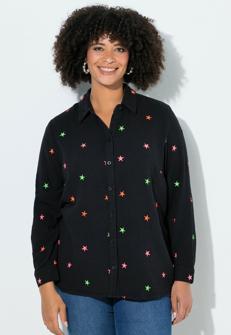 Vrouw met krullend haar die lacht, met grote oorbellen, een zwart shirt met neonroze, oranje en groene sterren, en een blauwe spijkerbroek, tegen een witte achtergrond.