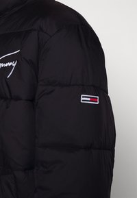 Czarna kurtka puchowa z haftowanym wzorem. Posiada biały logo "Tommy" na klatce piersiowej i kolorowy emblemat na rękawie. Gładka, błyszcząca struktura.