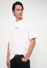 Mann mit dunklen Haaren, der ein lichtrosa T-Shirt von Calvin Klein und eine dunkelblaue Jeans trägt, steht mit einer Hand in der Tasche vor einem weißen Hintergrund.