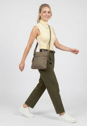 Junge Frau beim Gehen, trägt ein ärmelloses cremefarbenes Rollkragenoberteil, olivgrüne Hosen, weiße Sneakers und eine olivgrüne Umhängetasche.