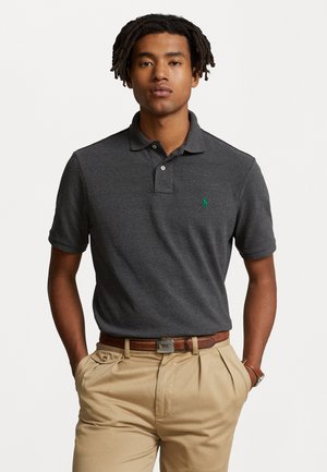 Polo shirt grigio scuro a maniche corte con una patta a tre bottoni e un logo ricamato verde sul lato sinistro del petto. Indossato con pantaloni beige chiaro.