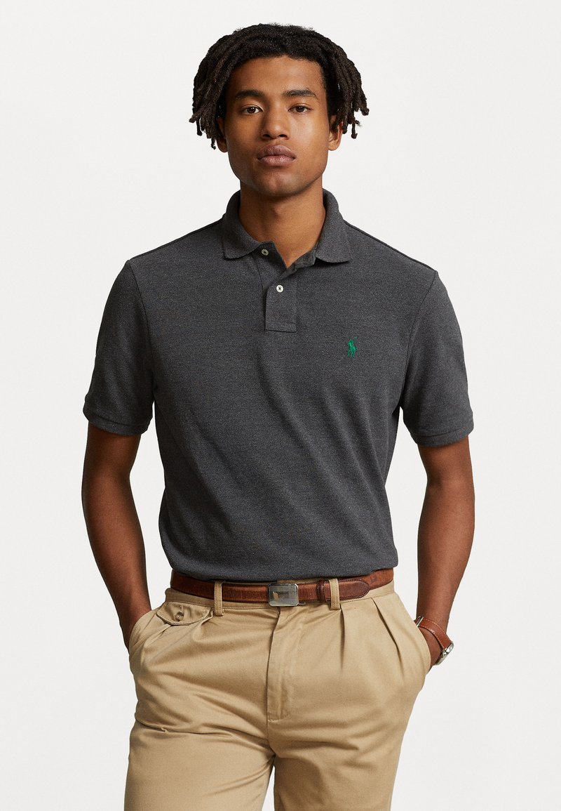 Polo de manga corta en gris oscuro con un tapeta de tres botones y un logo bordado verde en el lado izquierdo del pecho. Combinado con pantalones beige claros.