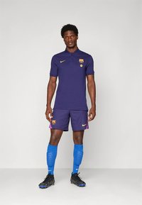 Mørklilla fotballdrakt med en polotrøye og shorts, med en klubblogo og gule detaljer. Sorte fotballsko og blå sokker fullfører antrekket.